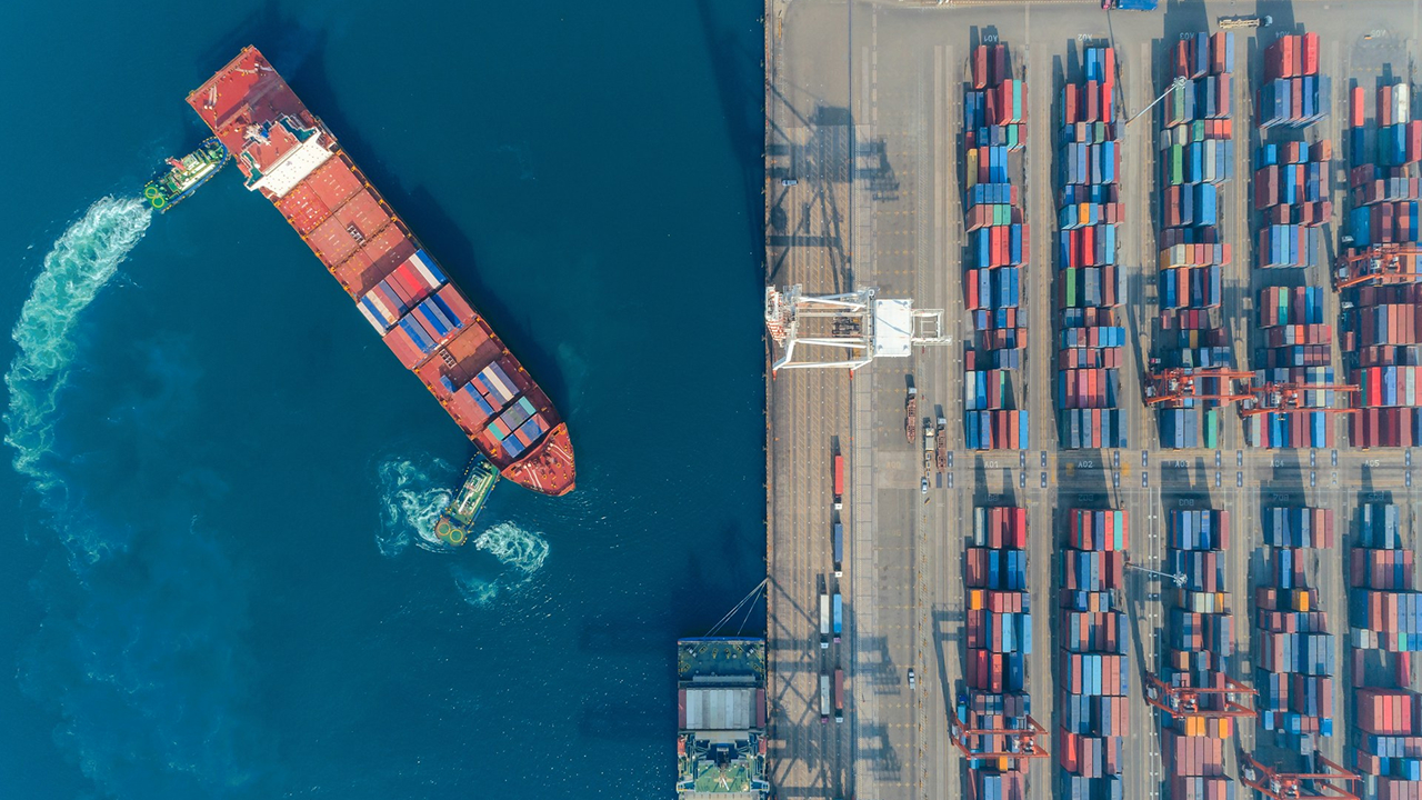 Vista aérea de um porto com um navio cargueiro manobrando para atracar, acompanhado de rebocadores, e diversos contêineres alinhados no terminal.