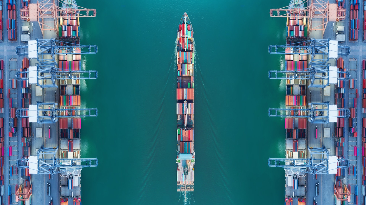 Navio cargueiro visto de cima navegando entre guindastes portuários e fileiras de contêineres organizados no terminal.